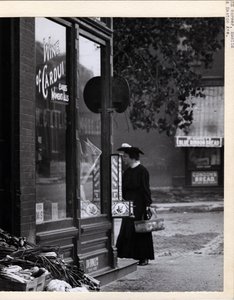 Lodret, sort og hvidt fotografi, der viser en kvinde, der holder en kurv, mens hun stod foran W. C. Kalis Drugstore -indgangen på 4878 Easton Avenue